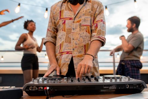 Deejay in shirt mixing sounds while young people dancing on background at rooftop party